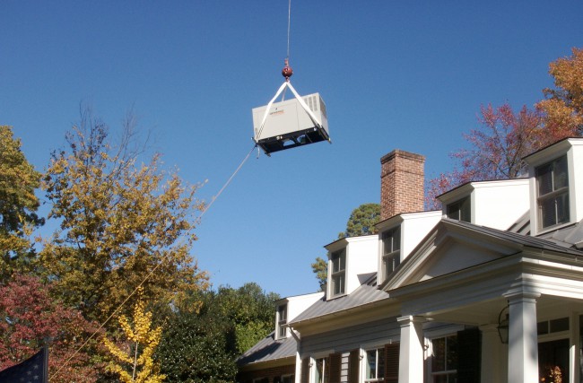 A Standby generator is being hoisted over a home by a crane.